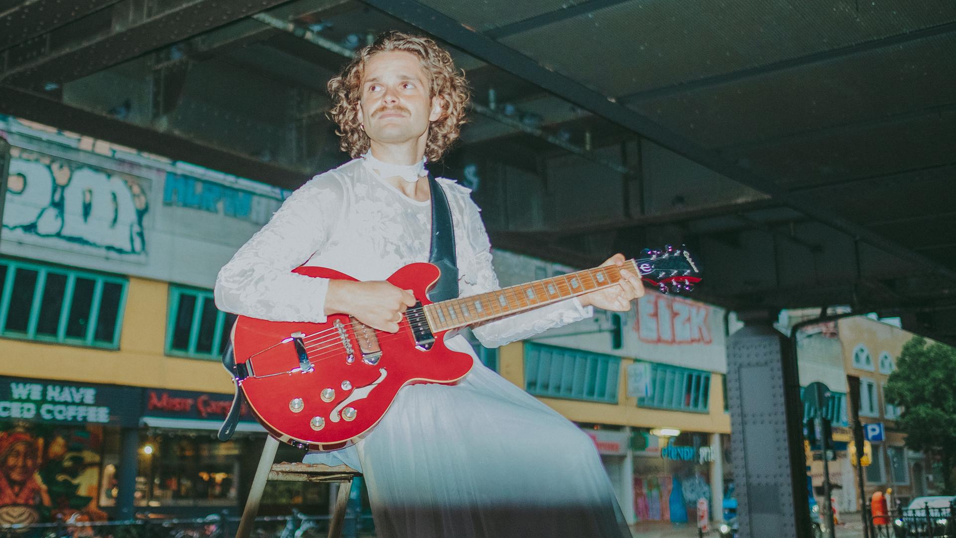 Tristan Brusch sitzt unter einer Berliner U-Bahn-Trasse und spielt eine knallrote E-Gitarre. Tristan Brusch sitzt unter einer Berliner U-Bahn-Trasse und spielt eine knallrote E-Gitarre.