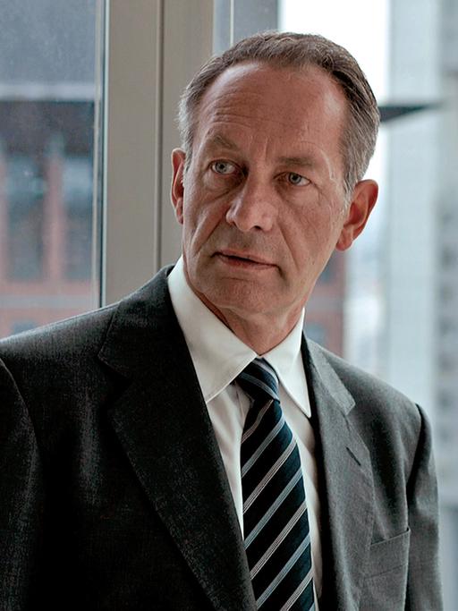 Der Schauspieler Robert Hunger-Bühler steht im Anzug in einem Büro. Durch die Glasfenster sieht man die Hochhäuser von Frankfurt.
