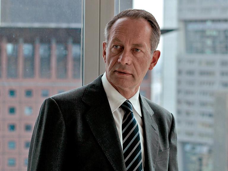 Der Schauspieler Robert Hunger-Bühler steht im Anzug in einem Büro. Durch die Glasfenster sieht man die Hochhäuser von Frankfurt.