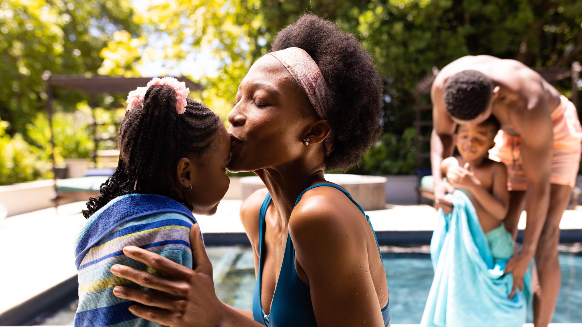 Eine Mutter küsst ihre Tochter auf die Stirn, während im Hintergrund der Vater seinen Sohn am Pool mit dem Handtuch abtrocknet. Eine Mutter küsst ihre Tochter auf die Stirn, während im Hintergrund der Vater seinen Sohn am Pool mit dem Handtuch abtrocknet.