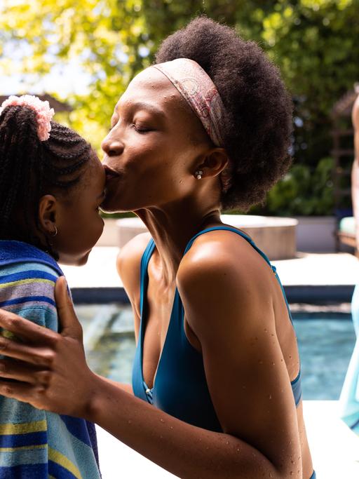 Eine Mutter küsst ihre Tochter auf die Stirn, während im Hintergrund der Vater seinen Sohn am Pool mit dem Handtuch abtrocknet. 