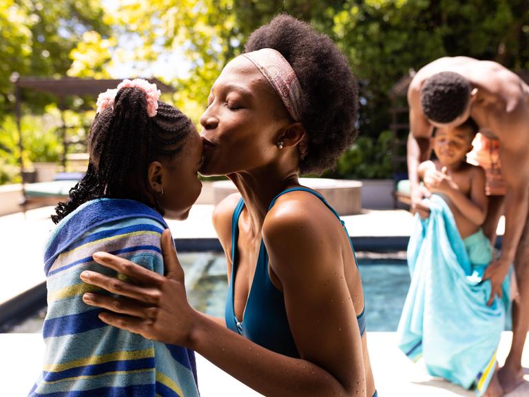 Eine Mutter küsst ihre Tochter auf die Stirn, während im Hintergrund der Vater seinen Sohn am Pool mit dem Handtuch abtrocknet. 