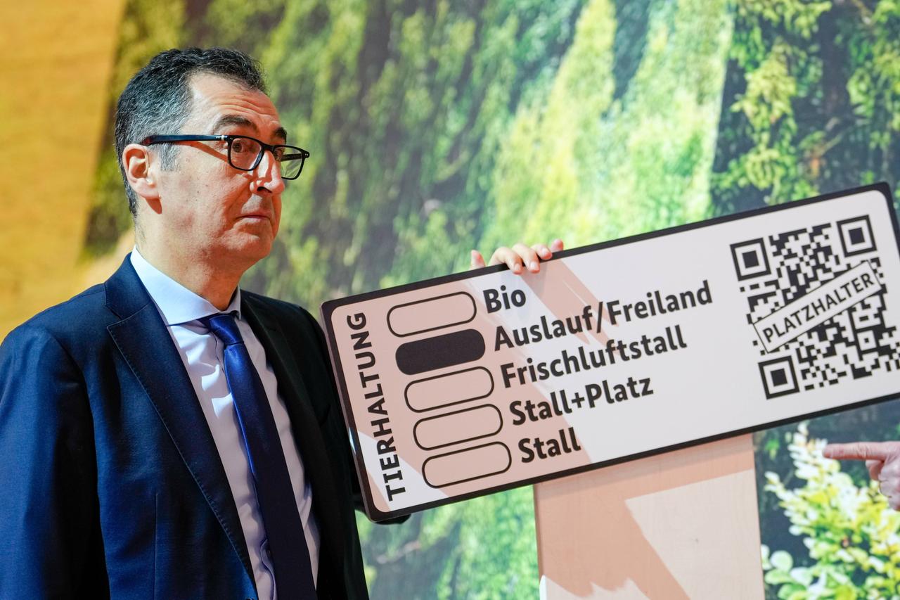 Cem Özdemir, ein Mann Mitte 50, hält eine Tafel hoch: Senkrecht steht auf dieser "Tierhaltung", dann folgen untereinander fünf Kategorien mit einem Kästschen davor: Bio, Auslauf/Freiland, Frischluftstall, Stall+ Platz sowie Stall, das Kästchen Auslauf/Freiland ist durch eine schwarze Füllung markiert. Ganz rechts ist ein QR-Code zu sehen.