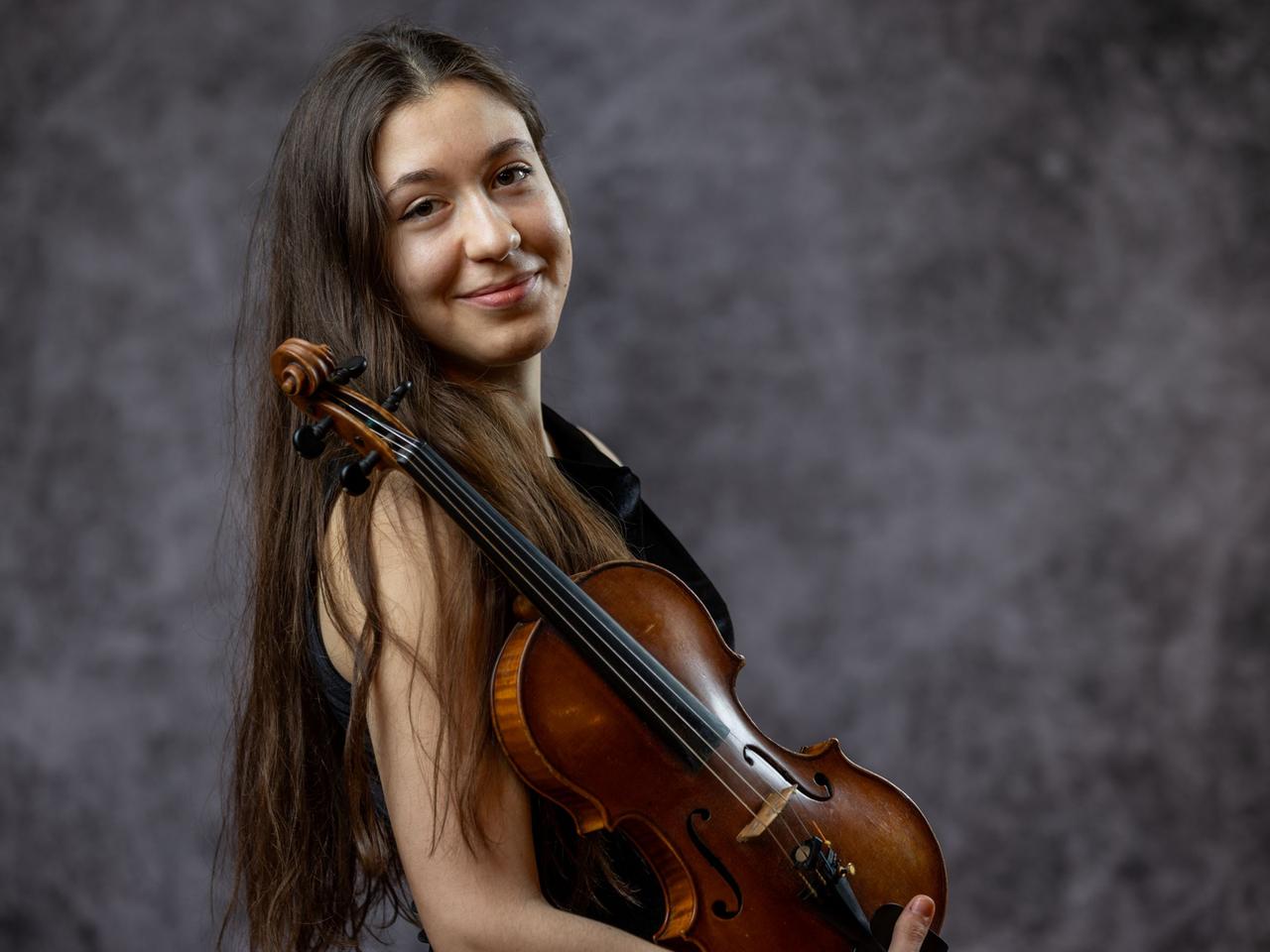 Dana Bostedt lacht mit ihren langen, braunen Haaren seitlich in die Kamera. Ihre Geige lehnt dabei auf ihrem Arm, gut sichtbar an ihre Schulter gelehnt.