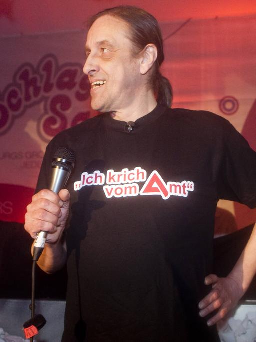 Arno Dübel steht mit einem Mikrofon in der Hand auf der Bühne. Auf seinem T-Shirt steht "Ich krich vom Amt".