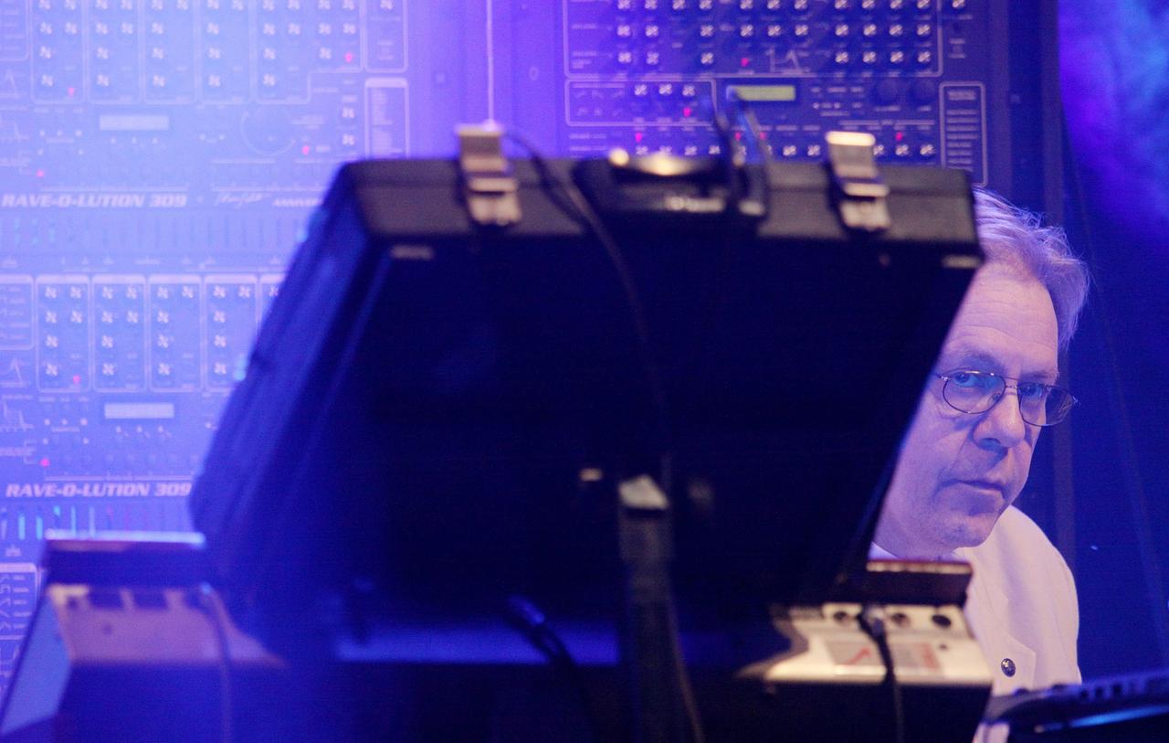 Klaus Schulze sitzt auf der Bühne und blickt hinter einem Synthesizer hervor und in die Kamera. Auch hinter ihm sind große Synthesizer-Wände zu sehen.