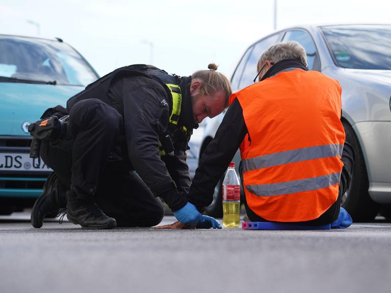 Ein Polizist versucht die Hand eines Klimaaktivisten vom Straßenbelag zu entfernen, der Aktivist Gruppe „Letzte Generation“ hat sich an auf diesen geklebt