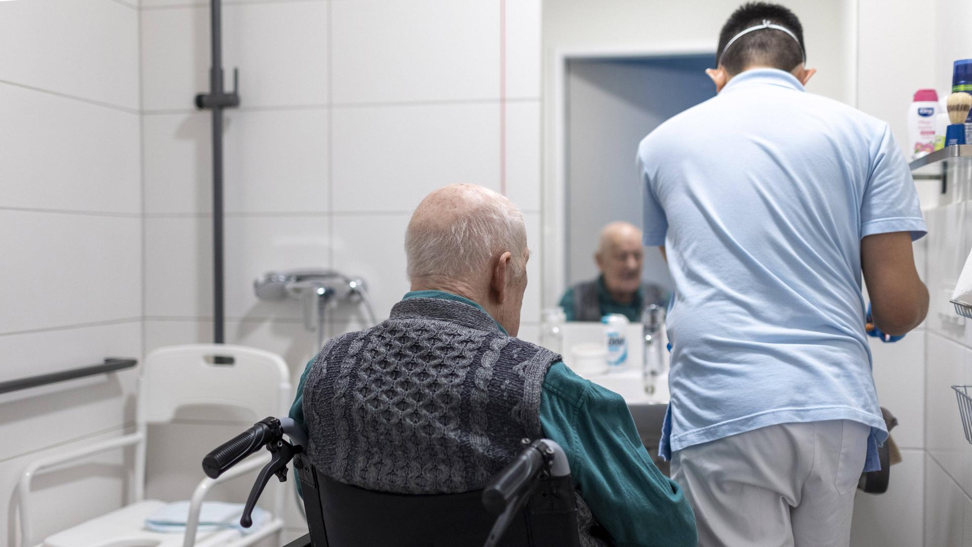 Ein Pfleger von hinten und ein alter Mann im Rollstuhl im Badezimmer eines Altersheims. 