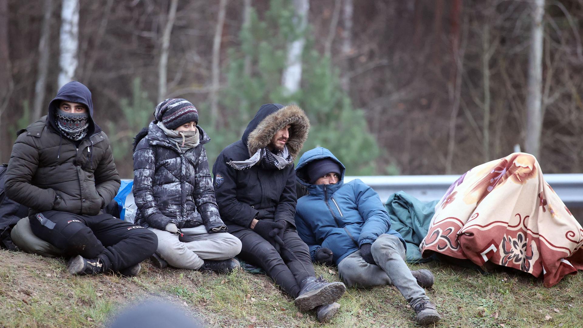 Vier Flüchtlinge, alle in dicke Winterjacken gehüllt, sitzen draußen auf dem Boden, hinter ihnen befinden sich Bäume. 