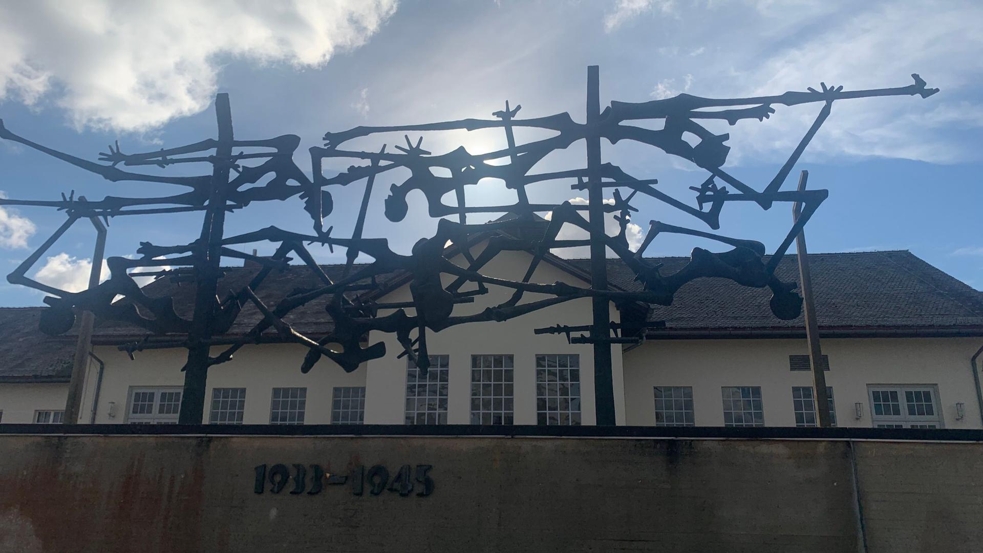 Blick von unten über einen Zaun mit Stahlgeflecht und Jahreszahlen auf dem Gelände des ehemaligen KZs Dachau. Zu sehen ist eines der Gebäude.