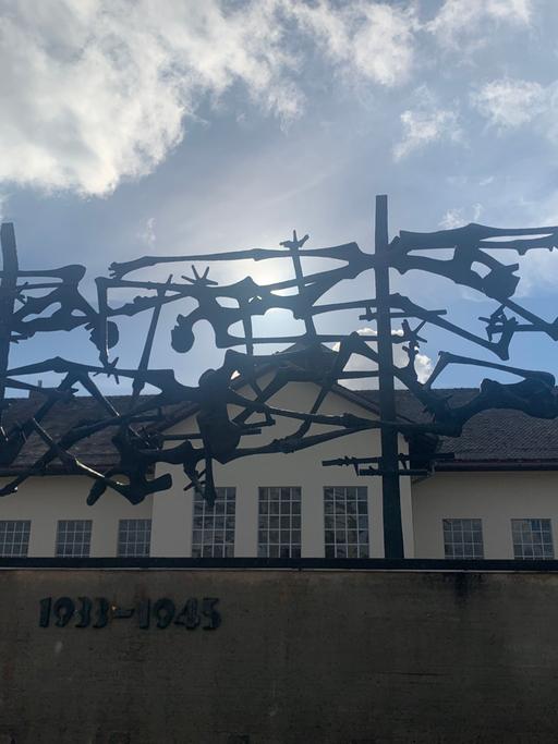Blick von unten über einen Zaun mit Stahlgeflecht und Jahreszahlen auf dem Gelände des ehemaligen KZs Dachau. Zu sehen ist eines der Gebäude.