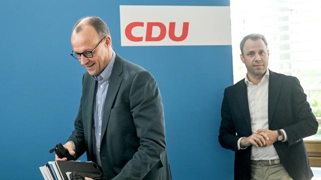 Zwei Männer mit Akten in der Hand und in Anzügen setzen sich an einen Tisch. Im Hintergrund der Schriftzug "CDU".
