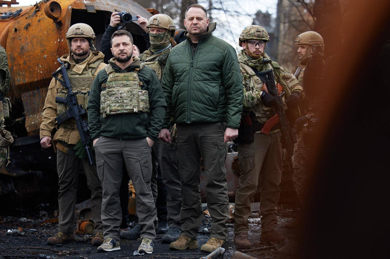 Der ukrainische Präsident Wolodymyr Selenskyj steht inmitten von Militärs und Begleitpersonen in zivil zwischen rostigen Trümmern.