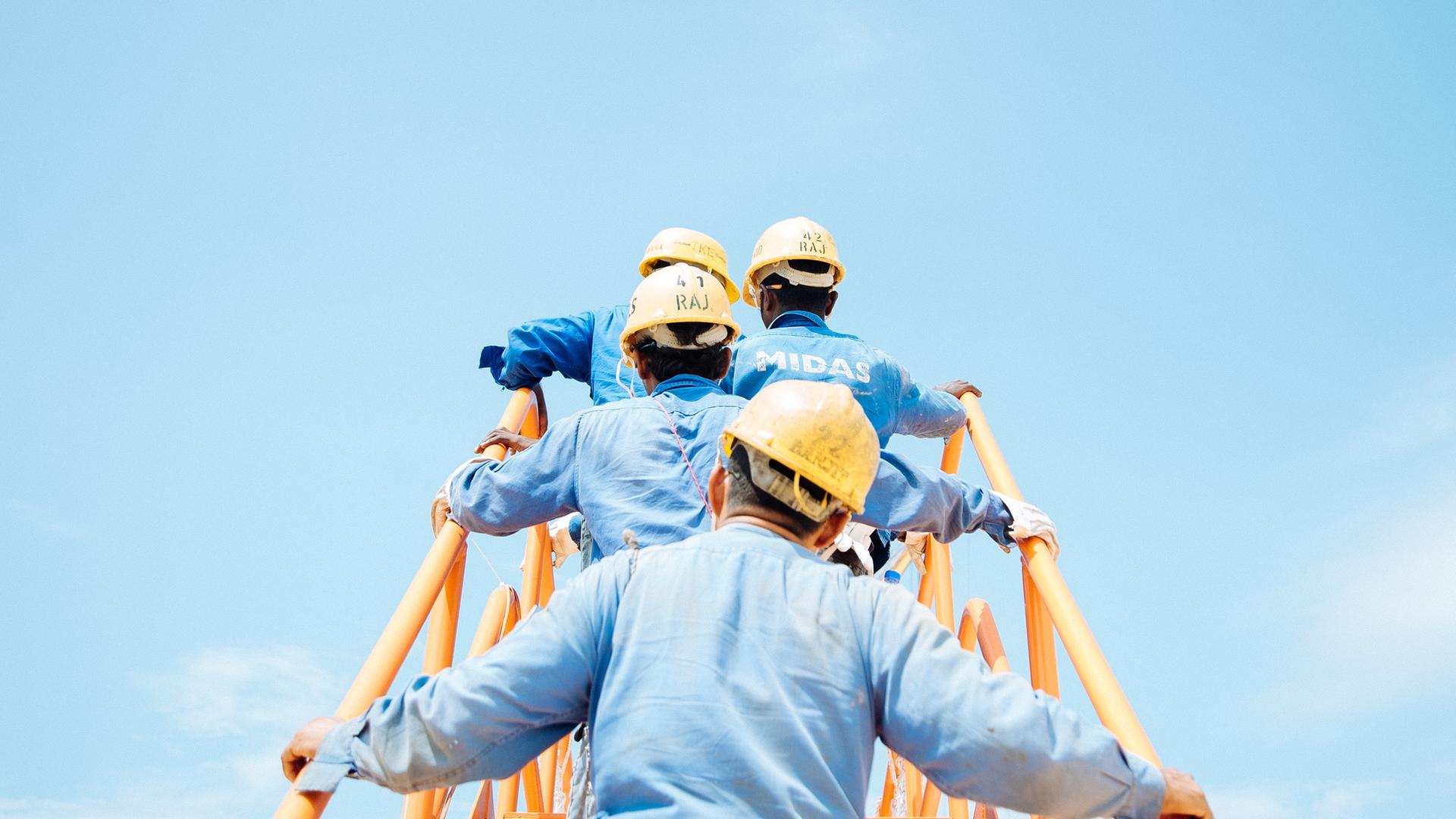 Eine Gruppe Männer in blauen Hemden und mit gelben Baustellenhelmen auf dem Kopf, gehen eine gelbe Metalltreppe hinauf. Rückansicht. Vor ihnen (Bildhintergrund): strahlend blauer Himmel.