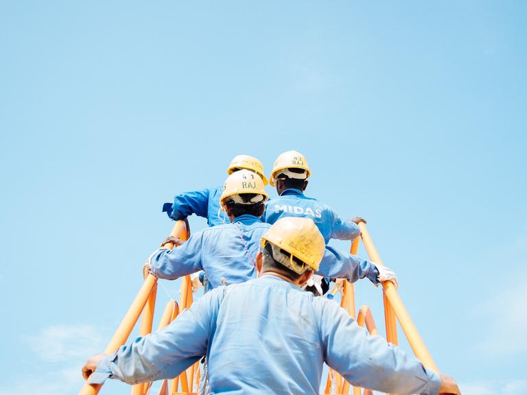 Eine Gruppe Männer in blauen Hemden und mit gelben Baustellenhelmen auf dem Kopf, gehen eine gelbe Metalltreppe hinauf. Rückansicht. Vor ihnen (Bildhintergrund): strahlend blauer Himmel.