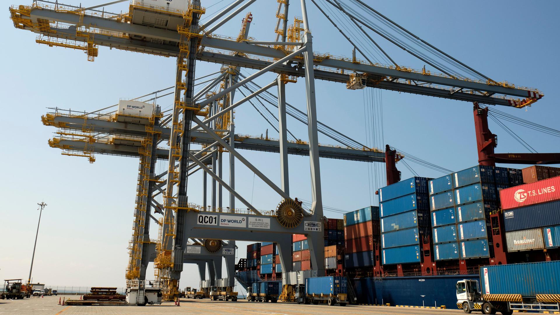 Große Kräne überragen die gestapelten blauen Container auf einem Schiff. Unten rechts kommt ein LKW angefahren. Große Kräne überragen die gestapelten blauen Container auf einem Schiff. Unten rechts kommt ein LKW angefahren.