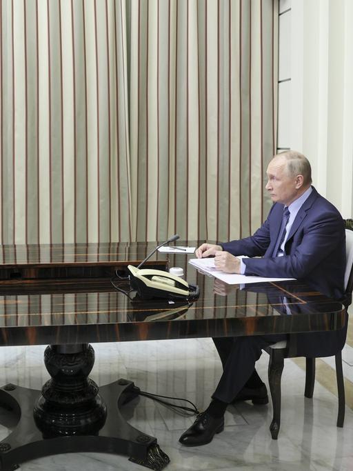 Der russische Präsident Vladimir Putin sitzt an einem langen, mächtigen Tisch und schaut auf einen Bildschirm, auf dem der amerikanische Präsident Joe Biden zu sehen ist.
