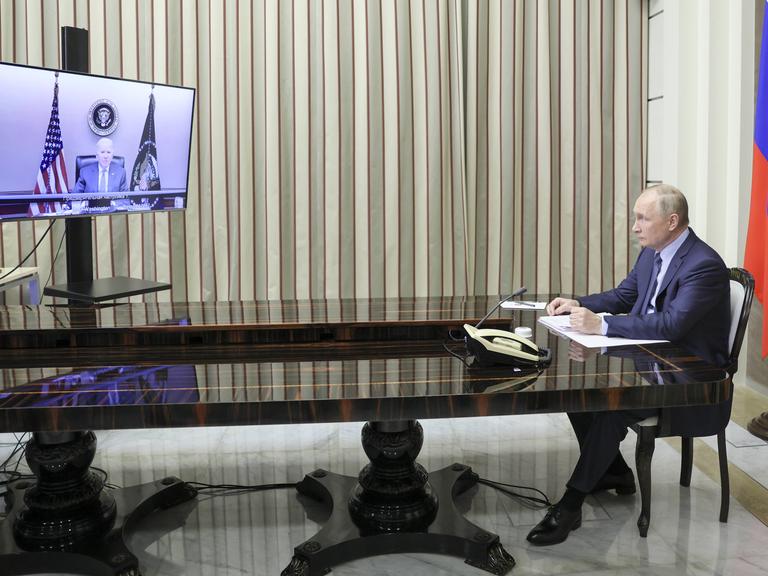 Der russische Präsident Vladimir Putin sitzt an einem langen, mächtigen Tisch und schaut auf einen Bildschirm, auf dem der amerikanische Präsident Joe Biden zu sehen ist.
