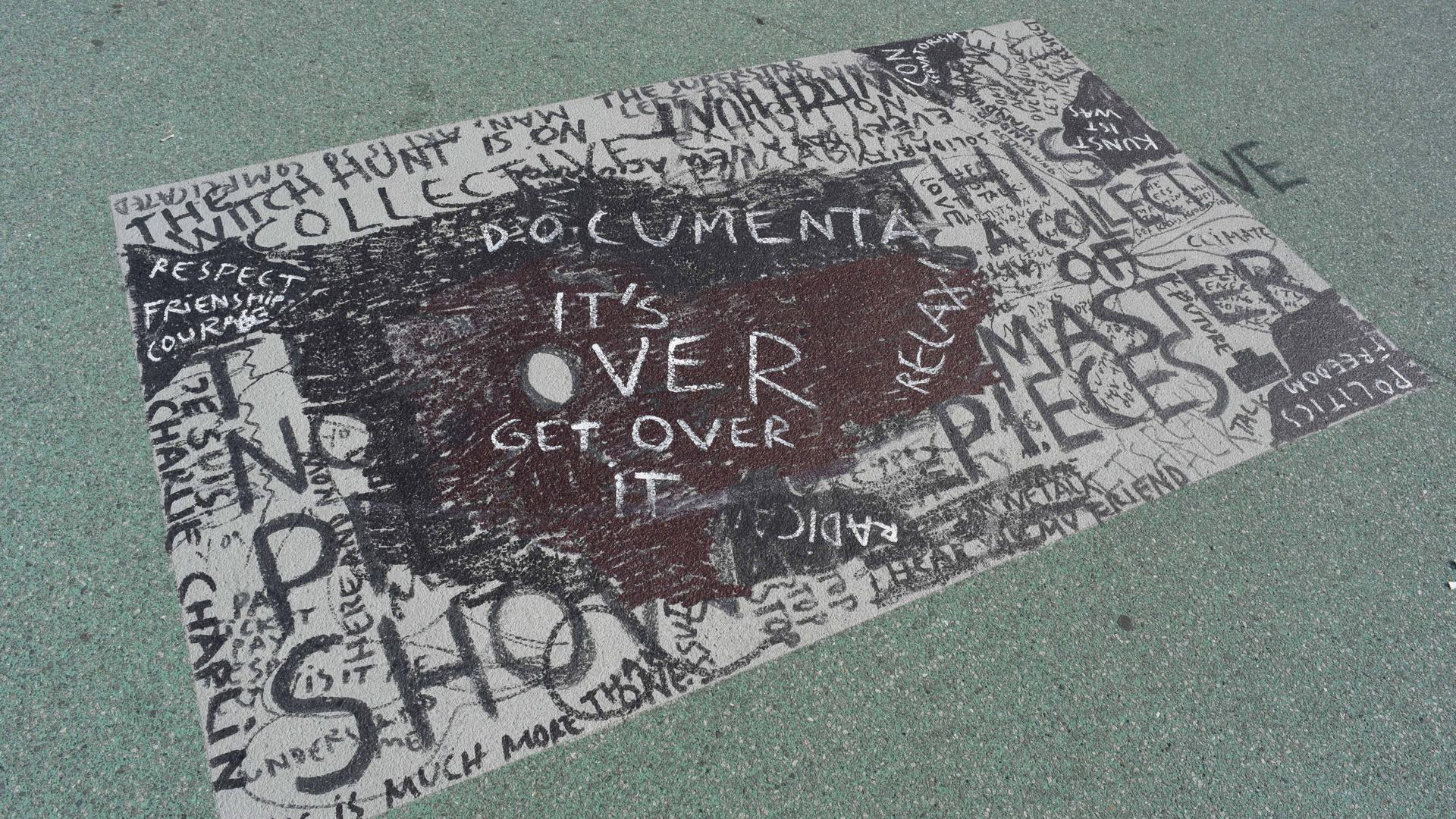 Ein Bodengemälde in dunklen Farben trägt den Schriftzug "Documenta. It's over. Get over it." Hinter und unter dem Hauptschriftzug sind weitere Worte zu erkennen. Ein Bodengemälde in dunklen Farben trägt den Schriftzug "Documenta. It's over. Get over it." Hinter und unter dem Hauptschriftzug sind weitere Worte zu erkennen.