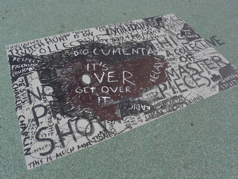 Ein Bodengemälde in dunklen Farben trägt den Schriftzug "Documenta. It's over. Get over it." Hinter und unter dem Hauptschriftzug sind weitere Worte zu erkennen.