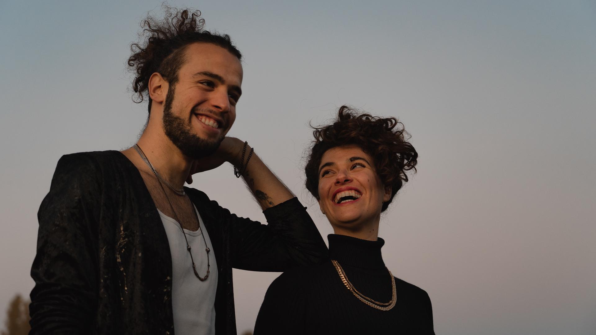 Ein junger Mann mit Zopf voller Locken und Bart stützt sich lachend mit seinem Ellenbogen auf die Schulter einer jungen Frau mit Locken, die ihn anschaut und dabei anlacht. Ein junger Mann mit Zopf voller Locken und Bart stützt sich lachend mit seinem Ellenbogen auf die Schulter einer jungen Frau mit Locken, die ihn anschaut und dabei anlacht.