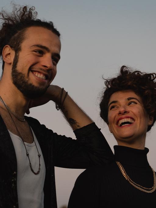 Ein junger Mann mit Zopf voller Locken und Bart stützt sich lachend mit seinem Ellenbogen auf die Schulter einer jungen Frau mit Locken, die ihn anschaut und dabei anlacht.