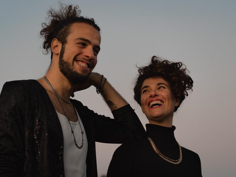 Ein junger Mann mit Zopf voller Locken und Bart stützt sich lachend mit seinem Ellenbogen auf die Schulter einer jungen Frau mit Locken, die ihn anschaut und dabei anlacht.
