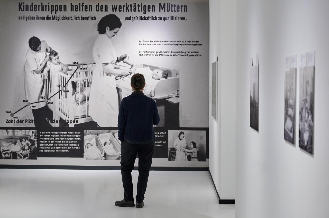Eine Person steht in der Rostocker Ausstellung vor einem Schaubild, das Betreuerinnen mit Kleinkindern zeigt. Darüber steht: "Kinderkrippen helfen den werktätigen Müttern." 