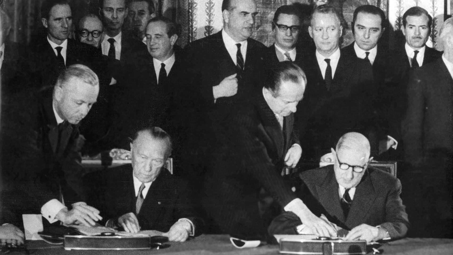 Ein schwarz weißes Foto vom 22. Januar 1963 : Zwei Männer sitzen nebeneinander an einem Tisch und unterzeichnen jeweils ein Dokument. Hinter ihnen stehen sehr viele Männer in Anzügen.