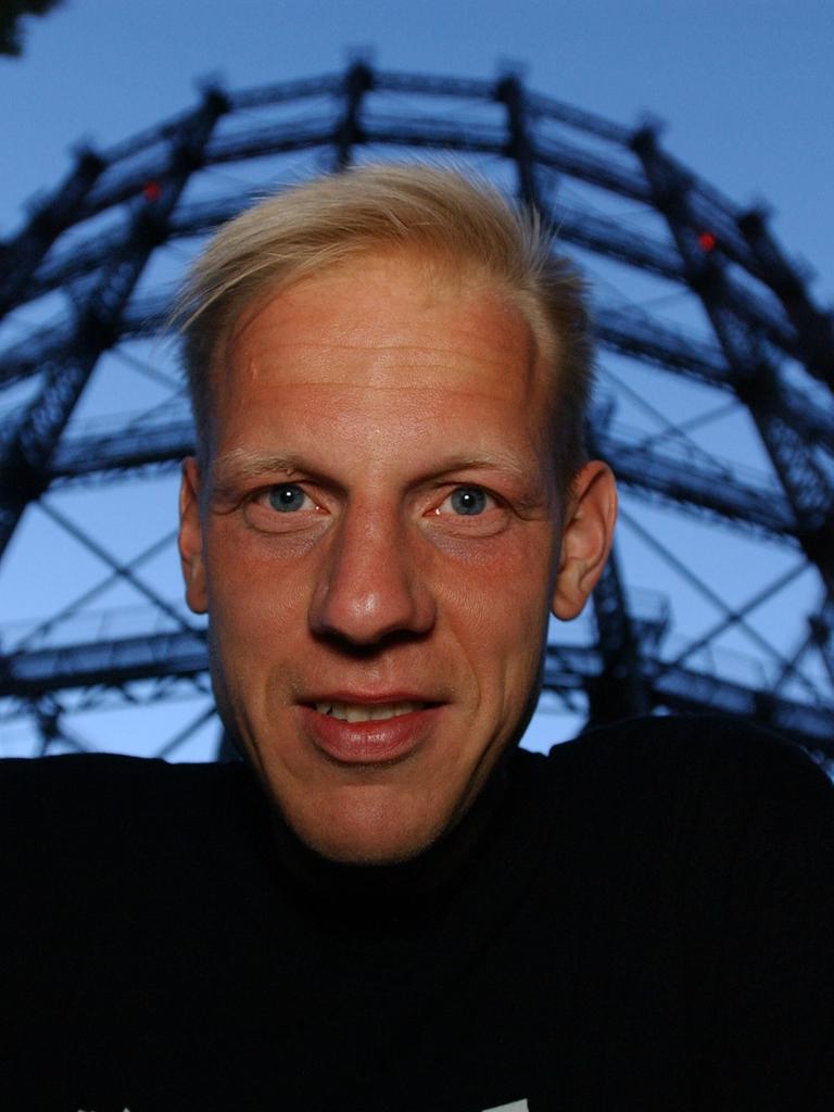 Der Filmemacher Jörg Buttgereit schaut in die Kamera, hinter ihm steht das ehemalige Gasometer in Berlin-Schöneberg.