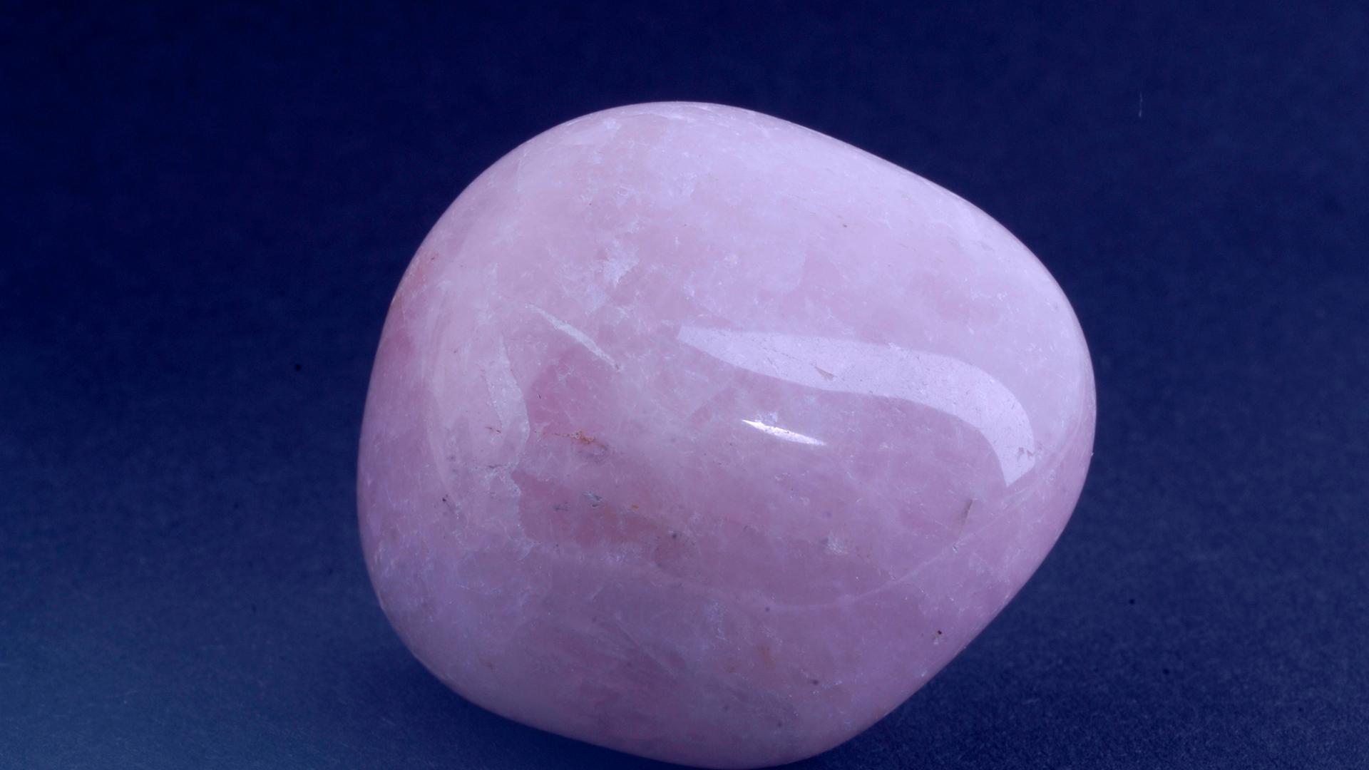 Ein geschliffener rosa Stein liegt auf einer dunkelblauen Fläche. Es ist ein Rosenquarz, der leicht weiß geadert ist. Seine Form ist rundlich. Ein geschliffener rosa Stein liegt auf einer dunkelblauen Fläche. Es ist ein Rosenquarz, der leicht weiß geadert ist. Seine Form ist rundlich.