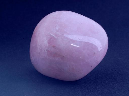 Ein geschliffener rosa Stein liegt auf einer dunkelblauen Fläche. Es ist ein Rosenquarz, der leicht weiß geadert ist. Seine Form ist rundlich.