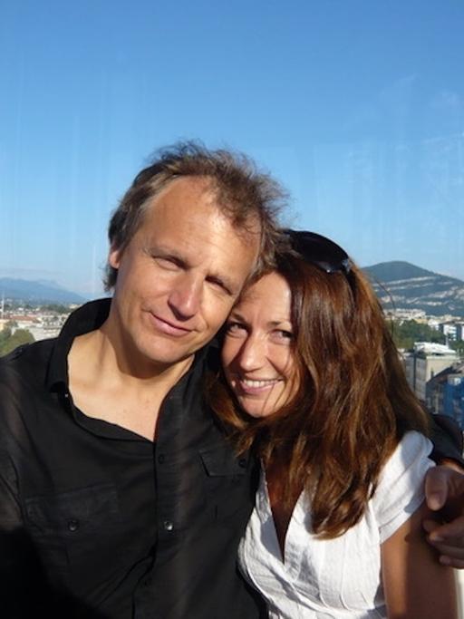 Ein Mann und eine Frau halten einander auf einem Balkon stehend in den Armen und lächeln in die Kamera.