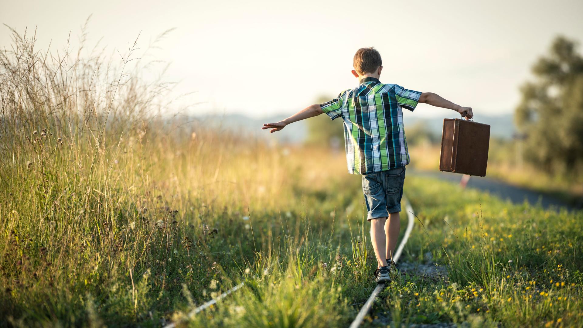Rückansicht eines Jungen, der einen Koffer in seinen ausgestreckten Armen hält. Er balanciert munter auf Schienen in der Abendsonne. Rückansicht eines Jungen, der einen Koffer in seinen ausgestreckten Armen hält. Er balanciert munter auf Schienen in der Abendsonne.