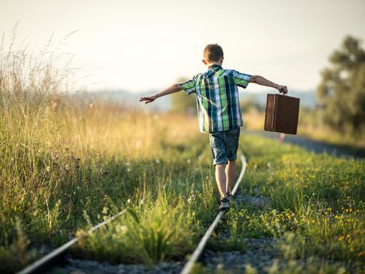 Rückansicht eines Jungen, der einen Koffer in seinen ausgestreckten Armen hält. Er balanciert munter auf Schienen in der Abendsonne.
