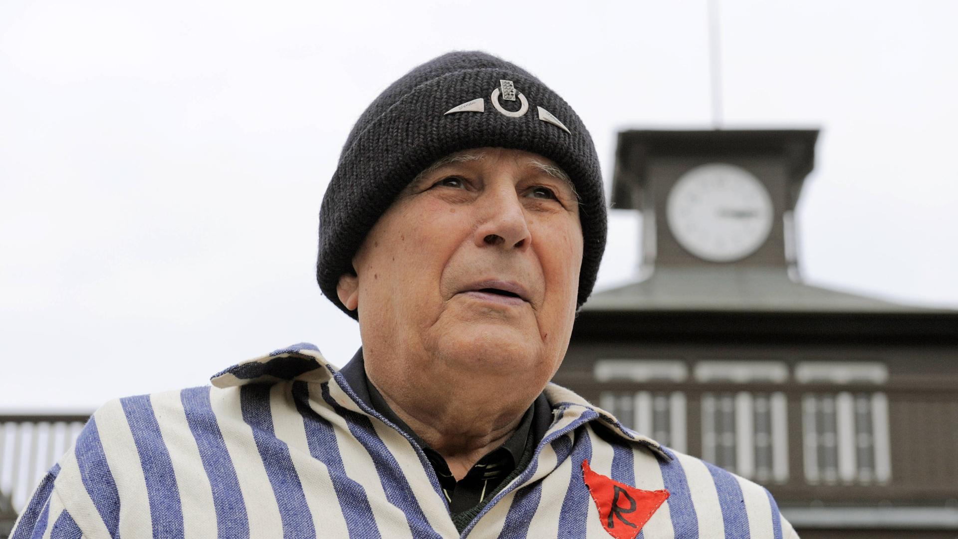 Ein Mann mit Wollmütze und KZ-Uniform steht vor dem Turm des KZs Buchenwald. Ein Mann mit Wollmütze und KZ-Uniform steht vor dem Turm des KZs Buchenwald.