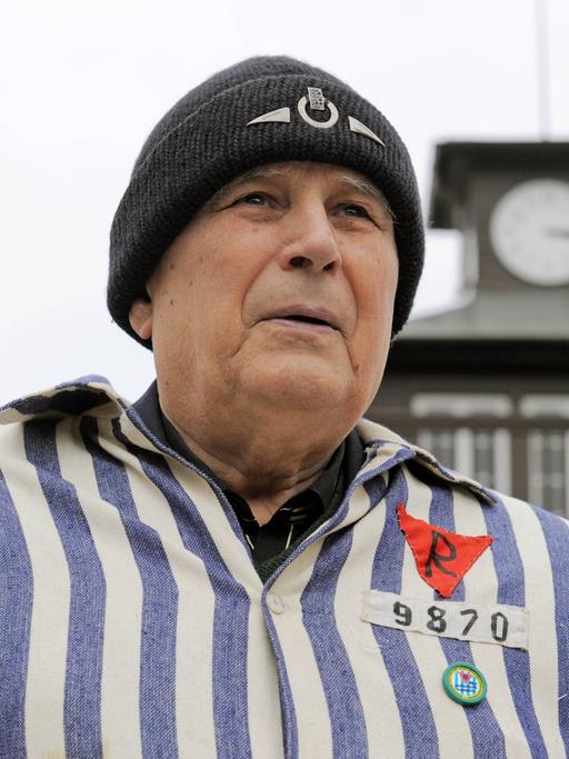 Ein Mann mit Wollmütze und KZ-Uniform steht vor dem Turm des KZs Buchenwald.