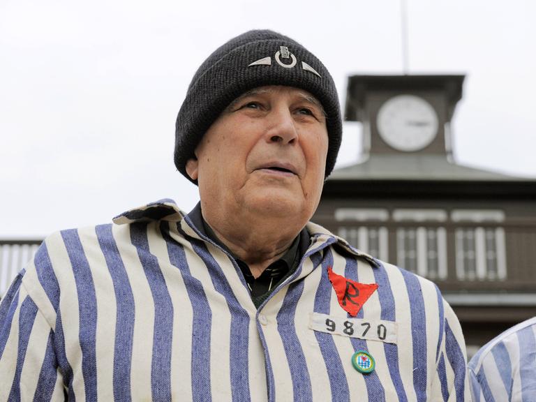Ein Mann mit Wollmütze und KZ-Uniform steht vor dem Turm des KZs Buchenwald.