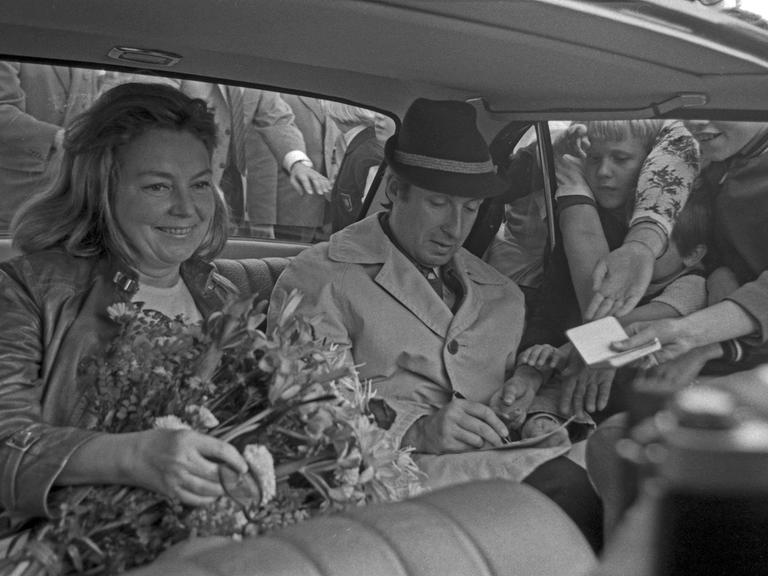 Schwarzweißbild von Peter Alexander und Hilde Haagen, die nebeneinander im Auto sitzen. Durch das Fenster stecken Fans Zettel mit Autogrammwünschen hinein.