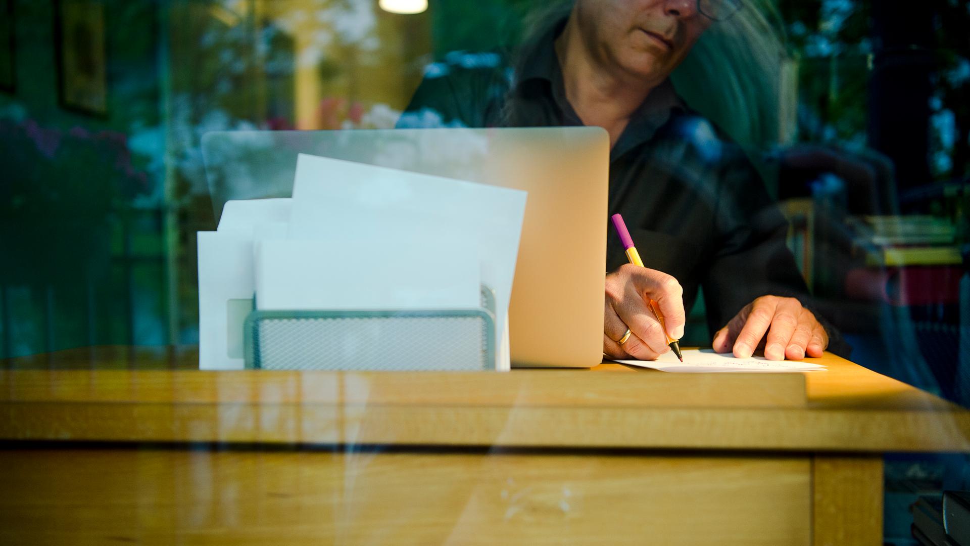 Ein Mann sitzt zu Hause an einem Schreibtisch. Er schreibt mit einem Stift auf ein Blatt Papier neben einem Laptop. Ein Mann sitzt zu Hause an einem Schreibtisch. Er schreibt mit einem Stift auf ein Blatt Papier neben einem Laptop.