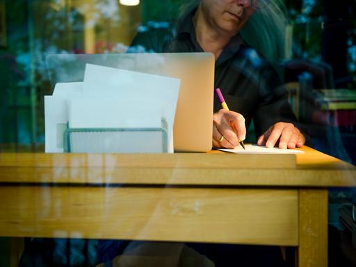 Ein Mann sitzt zu Hause an einem Schreibtisch. Er schreibt mit einem Stift auf ein Blatt Papier neben einem Laptop.