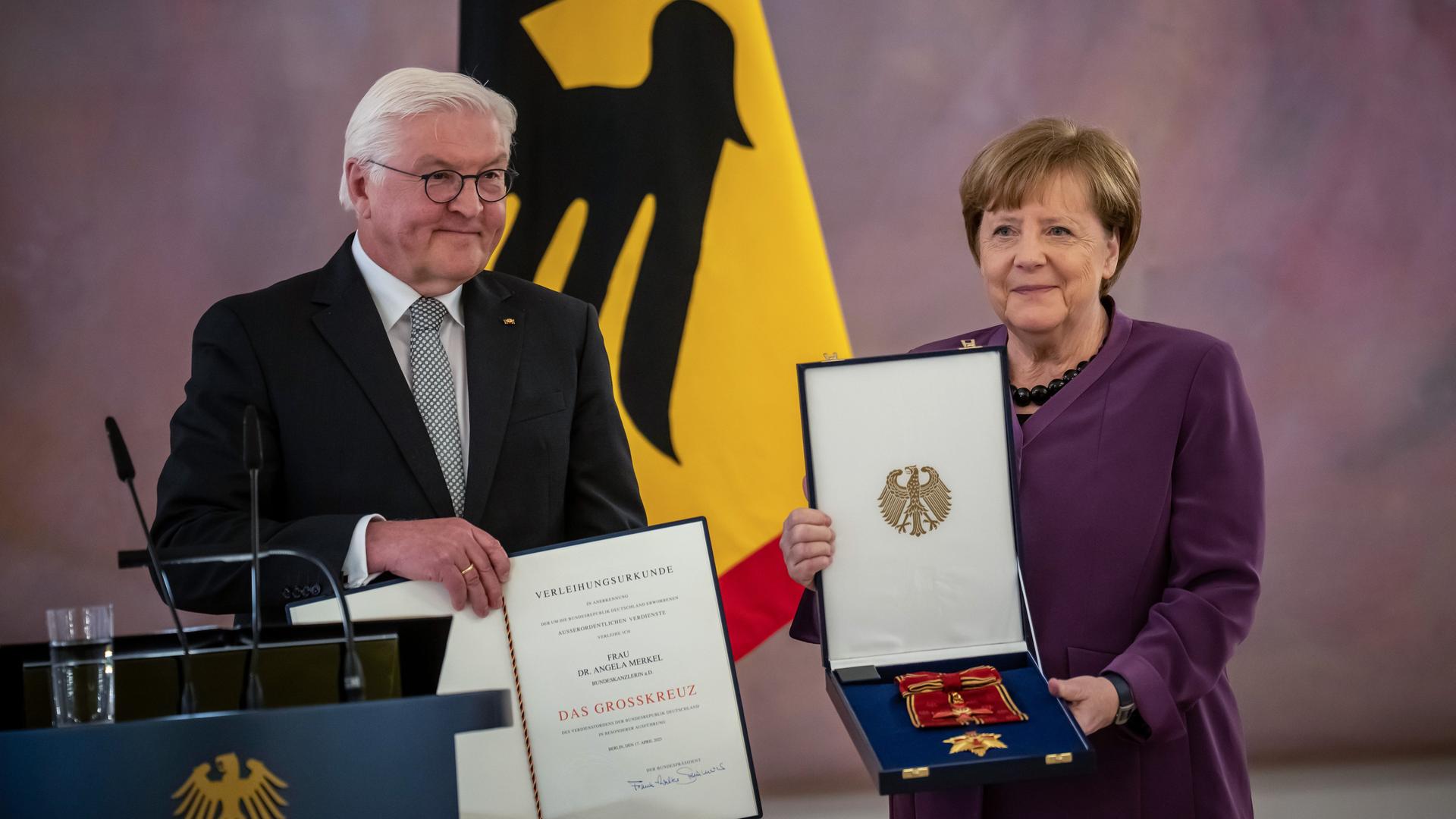 Die ehemalige Bundeskanzlerin Angela Merkel (CDU) steht neben Bundespräsident Frank-Walter Steinmeier und hält das Großkreuz des Verdienstordens der Bundesrepublik Deutschland in besonderer Ausführung mit beiden Händen vor sich.
