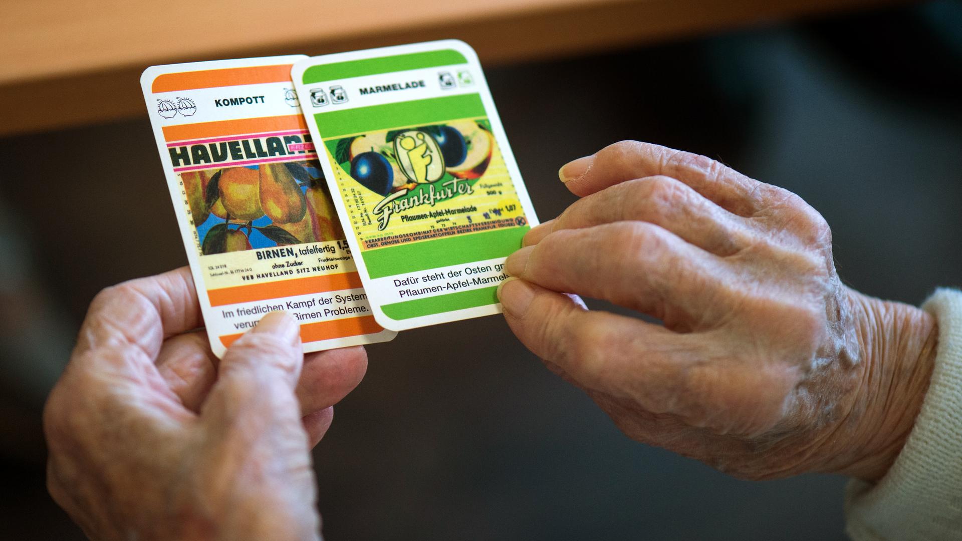 Zwei faltige Hände halten Spielkarten mit Fragen zu Produkten der DDR ("Kompott: Havelland"). Zwei faltige Hände halten Spielkarten mit Fragen zu Produkten der DDR ("Kompott: Havelland").