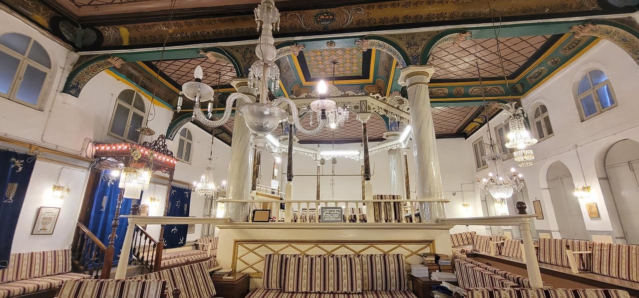 Im Innenraum einer Synagoge sind eine Kassettendecke mit hölzernen Ornamenten, mehrere weiße gläserne Kronleuchter und im Vordergrund zahlreiche gepolsterte Sitzbänke zu sehen.