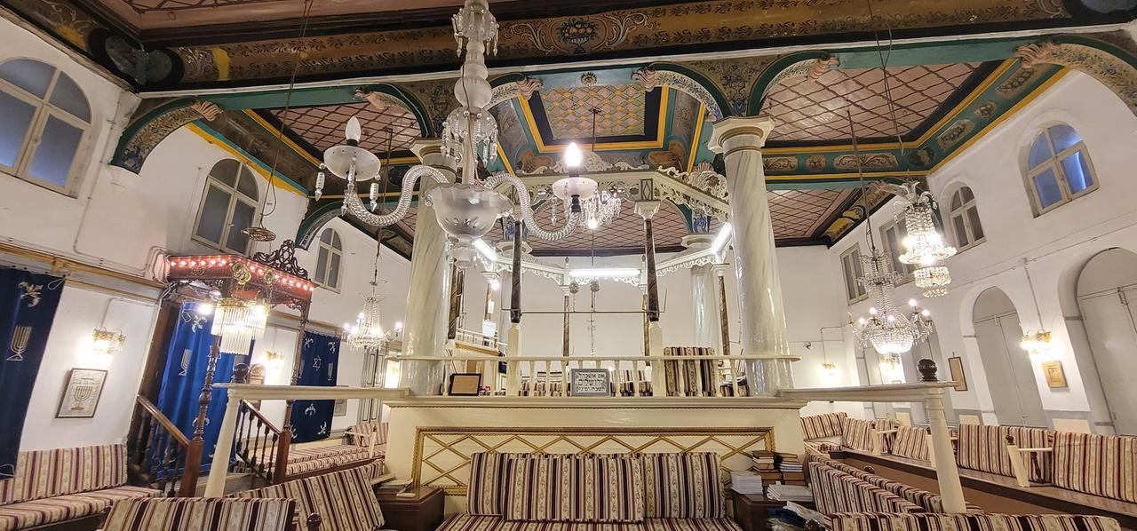 Im Innenraum einer Synagoge sind eine Kassettendecke mit hölzernen Ornamenten, mehrere weiße gläserne Kronleuchter und im Vordergrund zahlreiche gepolsterte Sitzbänke zu sehen. Im Innenraum einer Synagoge sind eine Kassettendecke mit hölzernen Ornamenten, mehrere weiße gläserne Kronleuchter und im Vordergrund zahlreiche gepolsterte Sitzbänke zu sehen.
