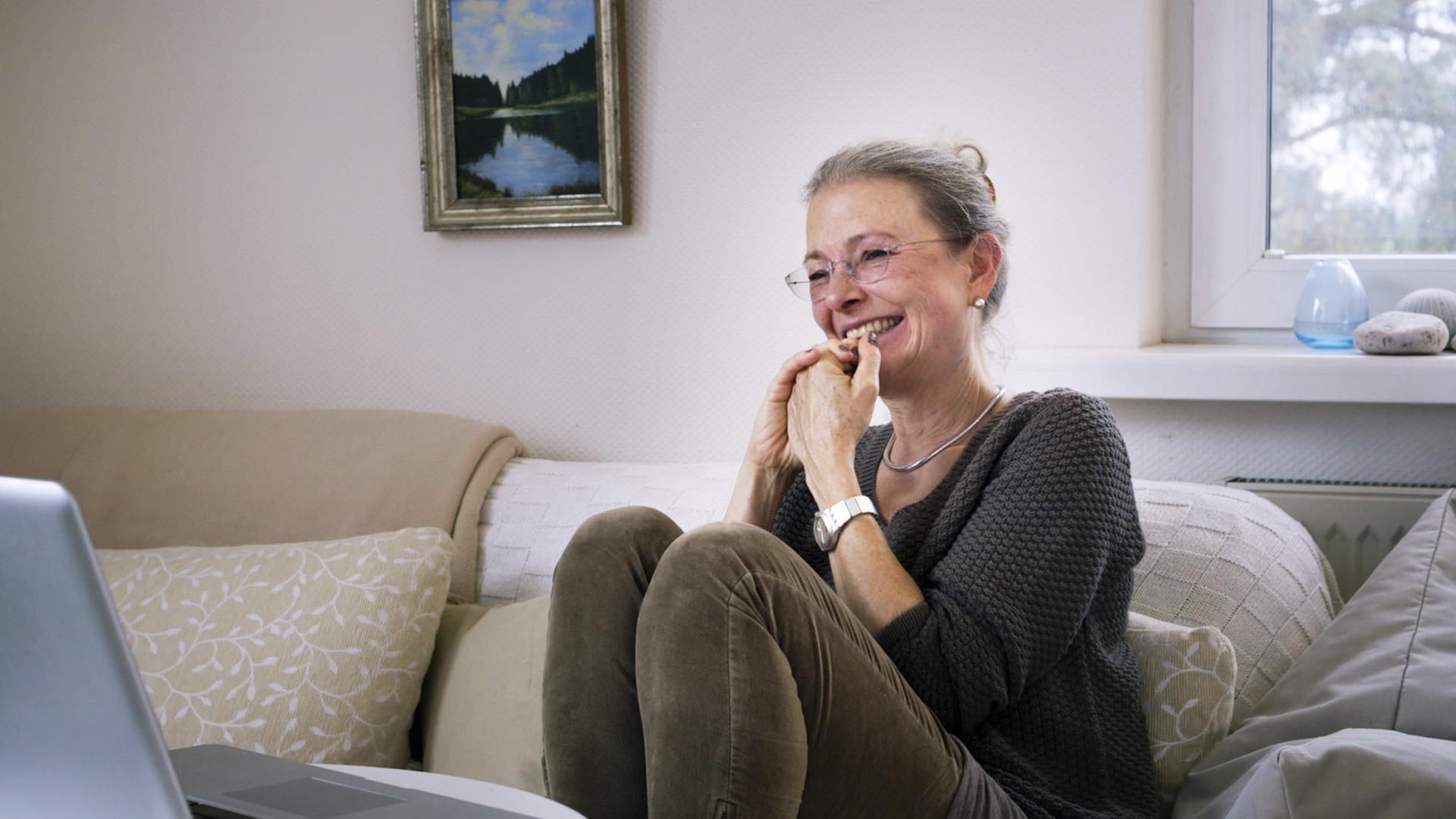 Filmstill aus der Doku: "ER SIE ICH" von Carlotta Kittel. Die Mutter sitzt auf einem Sofa und lacht. Filmstill aus der Doku: "ER SIE ICH" von Carlotta Kittel. Die Mutter sitzt auf einem Sofa und lacht.