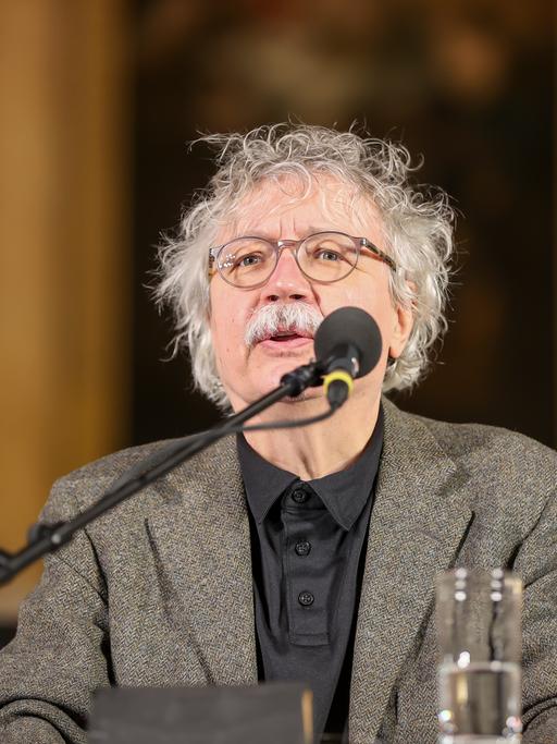 Ein Mann mit Brille, grauen Locken und Schnauzbart sitzt im grauen Jacket und schwarzem Hemd hinter einem Rednerpult und spricht in ein Mikrofon.