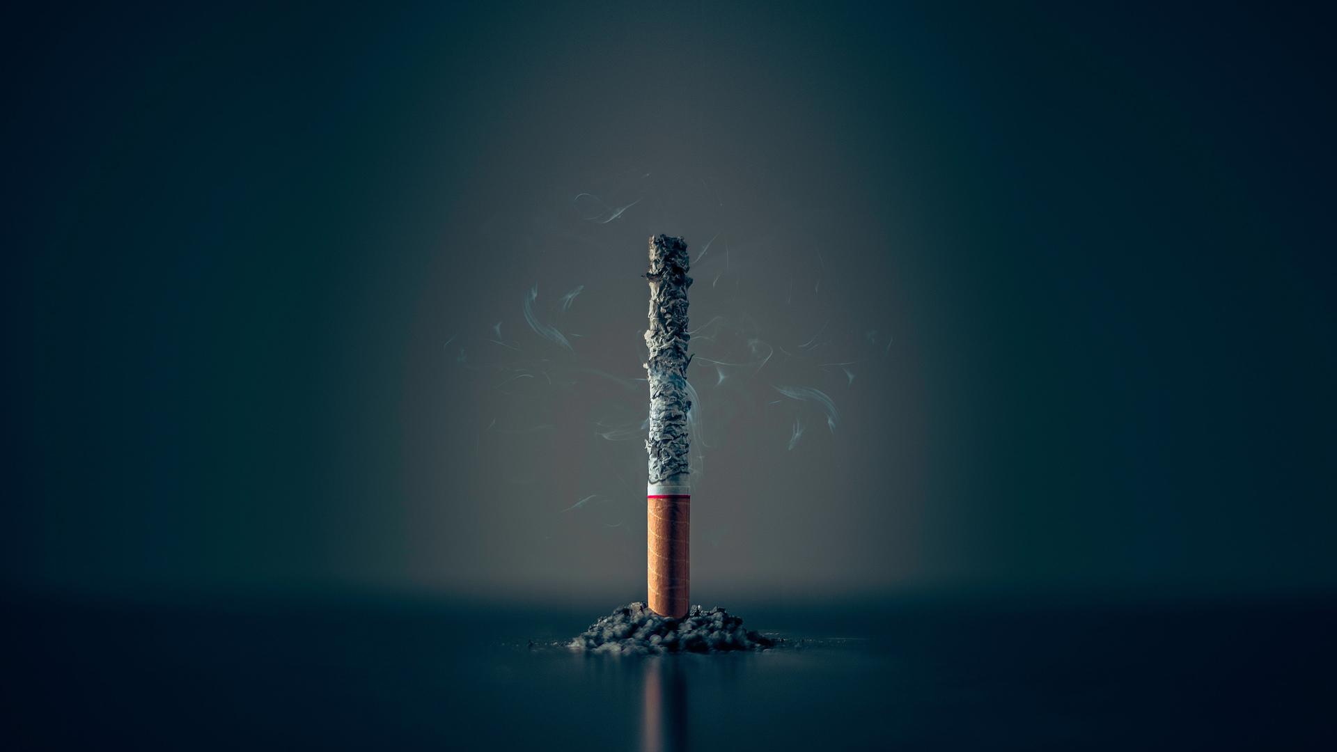 Eine einzelne Zigarette steht heruntergebrannt bis auf den Filter und noch qualmend aufrecht vor einem dunklen Hintergrund.