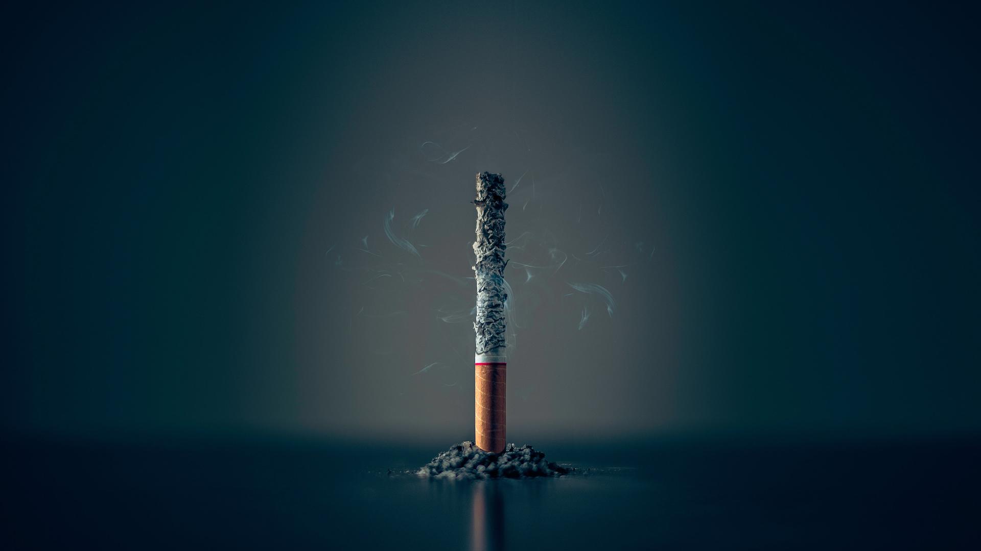 Eine einzelne Zigarette steht heruntergebrannt bis auf den Filter und noch qualmend aufrecht vor einem dunklen Hintergrund. Eine einzelne Zigarette steht heruntergebrannt bis auf den Filter und noch qualmend aufrecht vor einem dunklen Hintergrund.