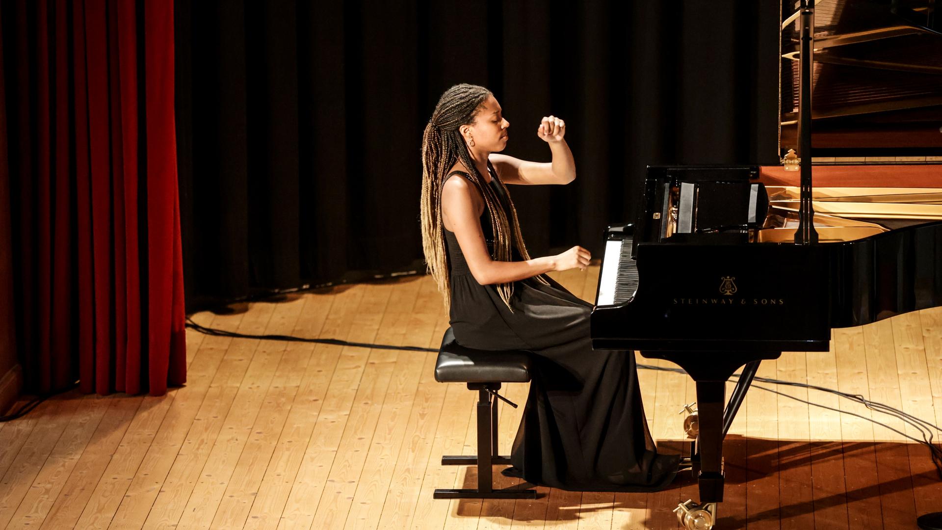 Eine Konzertbühne, darauf steht ein langer schwarzer Flügel. An den Tasten sitzt eine junge, schlanke Frau in einem schwarzen Kleid. Es ist die Pianistin Jeneba Kanneh-Mason.
